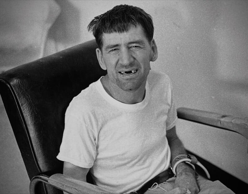 A man with short dark hair, wearing a white t-shirt, sits in a wheelchair and smiles. He appears to have a developmental disability. The image is in black and white.