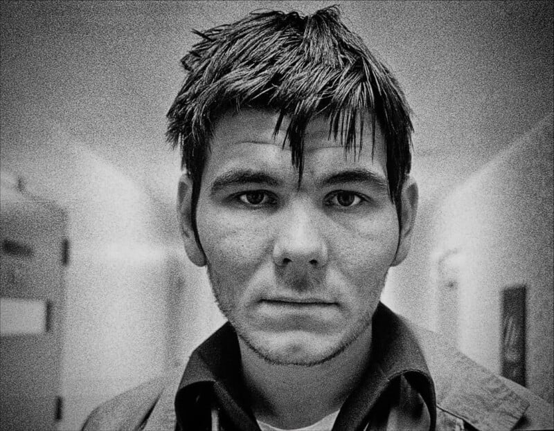 Black-and-white close-up photo of a young man with tousled dark hair and stubble, standing in a hallway with blank walls and a door visible in the background. He has a serious, contemplative expression.