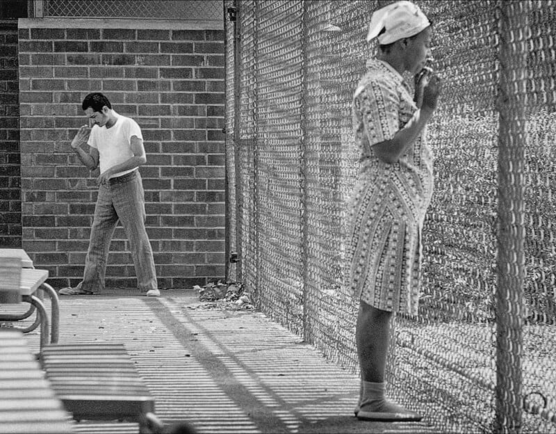 A man in a white shirt stands near a brick wall inside a fenced area, while a woman in a dress and headscarf stands on the other side of the fence, gazing through the wire mesh.