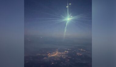 A bright green meteor blazes across the night sky, emitting glowing light with visible streaks, over a dark landscape dotted with city lights far below.
