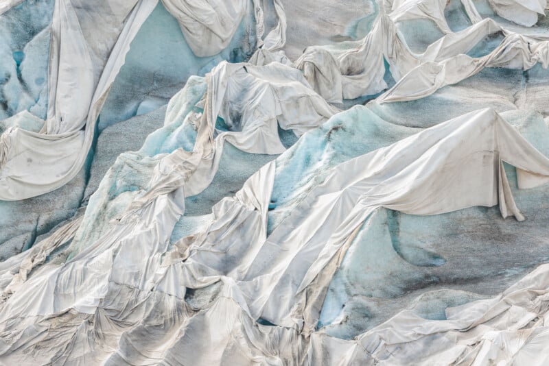 Large white sheets cover a blue and gray glacier, creating folds and creases over the ice to help slow melting. The fabric contrasts with the natural icy textures underneath.