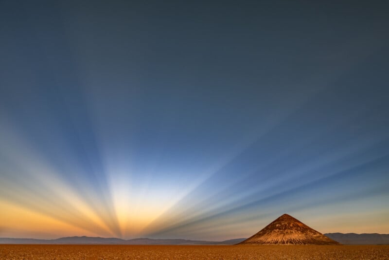 A small, conical hill stands on a flat desert plain under a clear sky at sunrise or sunset, with dramatic sun rays fanning outward and mountains visible in the distant background.