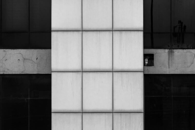 A tall building facade with large white panels in the center, flanked by dark windows. A small window on the right shows a person’s face peeking out. The image is in black and white, with a minimalistic, geometric composition.