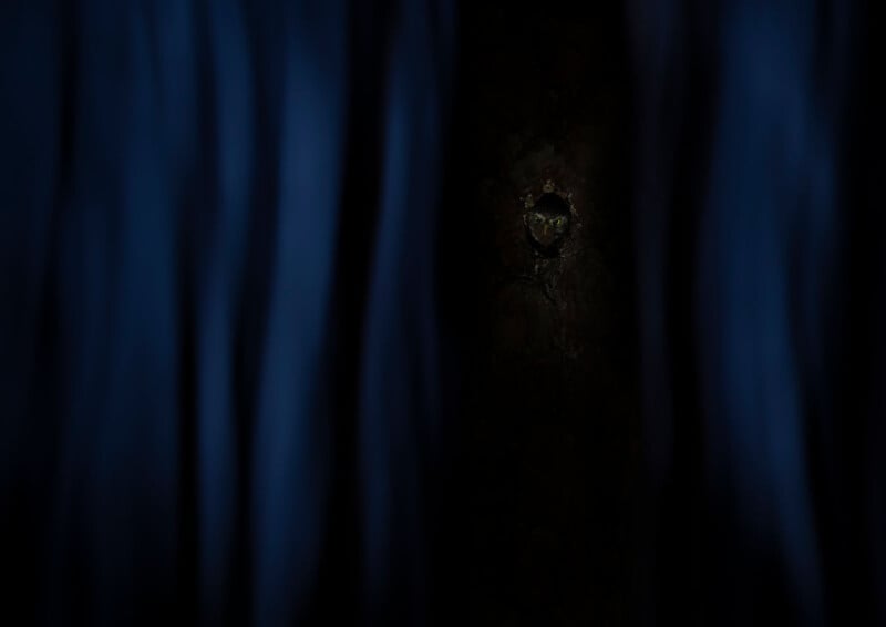 A single eye peers through a small hole in a dark wall, partially obscured by dark blue curtains on both sides, creating a mysterious and eerie atmosphere.