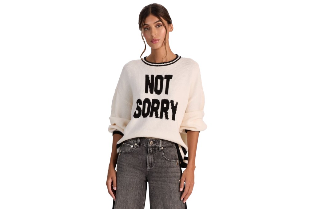 Woman wearing a cream sweater with "NOT SORRY" in black letters and black jeans.
