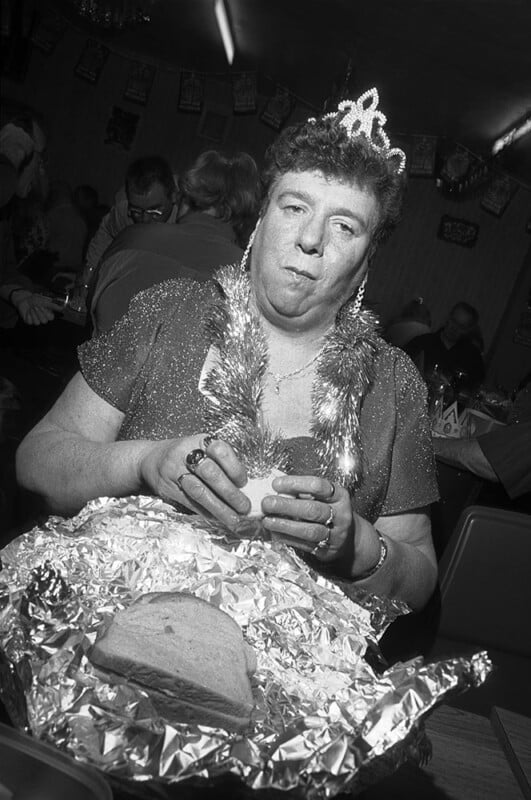 A person wearing a tiara, tinsel garland, and sparkly dress sits in a crowded room, holding a sandwich over foil-wrapped bread, looking thoughtful or pensive.