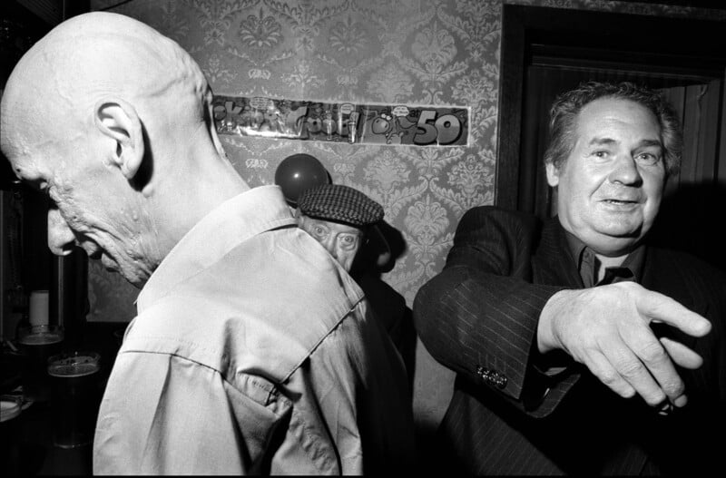 Three older men stand in a dimly lit room with patterned wallpaper; one faces away, one looks through a doorway, and another gestures with his arm. A “50” birthday banner and balloons are visible in the background.