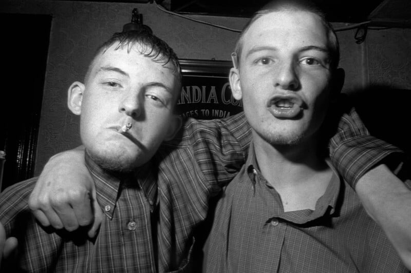 Two young men in plaid shirts, one with a cigarette in his mouth, pose closely together with arms around each other in a dimly lit room, appearing energetic and expressive.