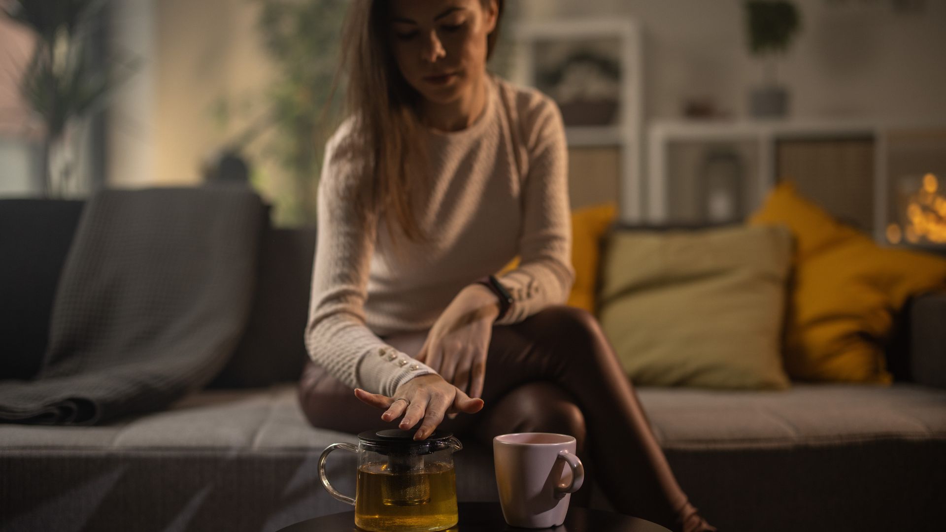 A woman sits on her couch with pot of chamomile tea and a cup on the table in front of her.