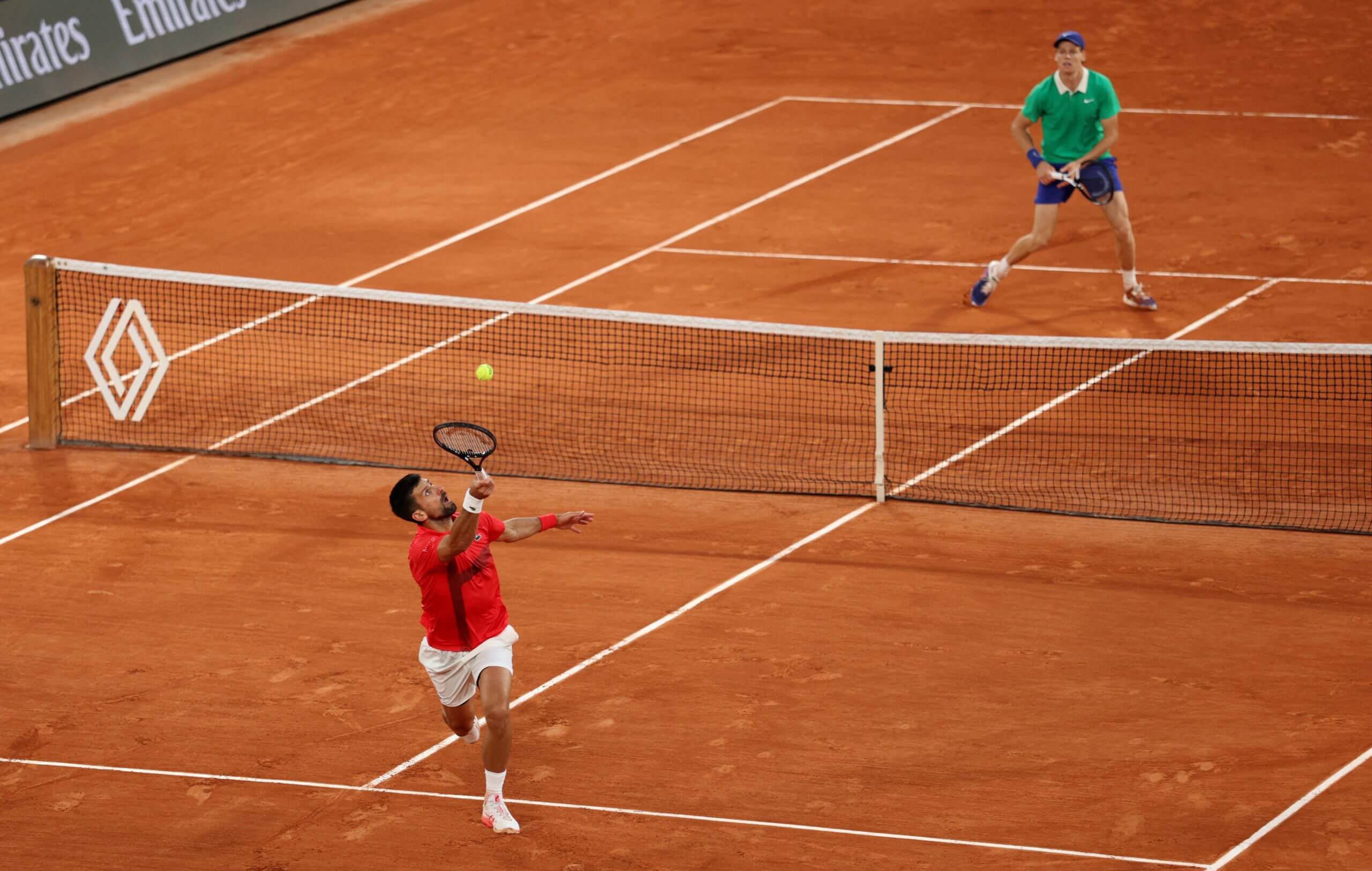Novak Djokovic stretches for a ball going over his head in the foreground on a clay tennis court, while Jannik Sinner waits in the background in front of the service line.