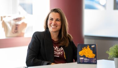 A woman in a blazer sits at a table, with a bag of coffee grounds on the table in front of her.