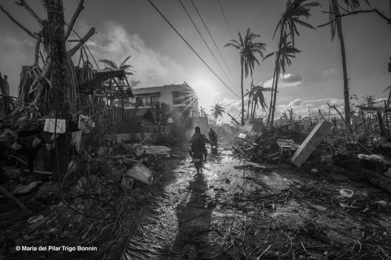 Two people on a motorbike ride through a muddy, debris-filled street lined with damaged trees and destroyed buildings after a disaster, as the sun sets behind them. The scene is desolate and filled with destruction.