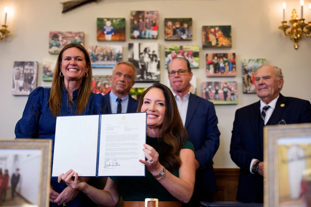 A group of officials stand behind a woman seated at a desk as she holds up a signed document. Framed photos hang on the wall behind them.