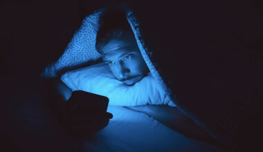 A person lies in bed under a blanket at night, staring at a smartphone that emits a blue glow, illuminating their face in the dark room.