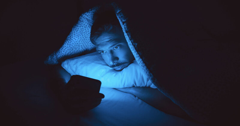 A person lies in bed under a blanket at night, staring at a smartphone that emits a blue glow, illuminating their face in the dark room.