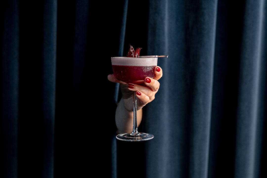 A person holds a cocktail through a curtain.