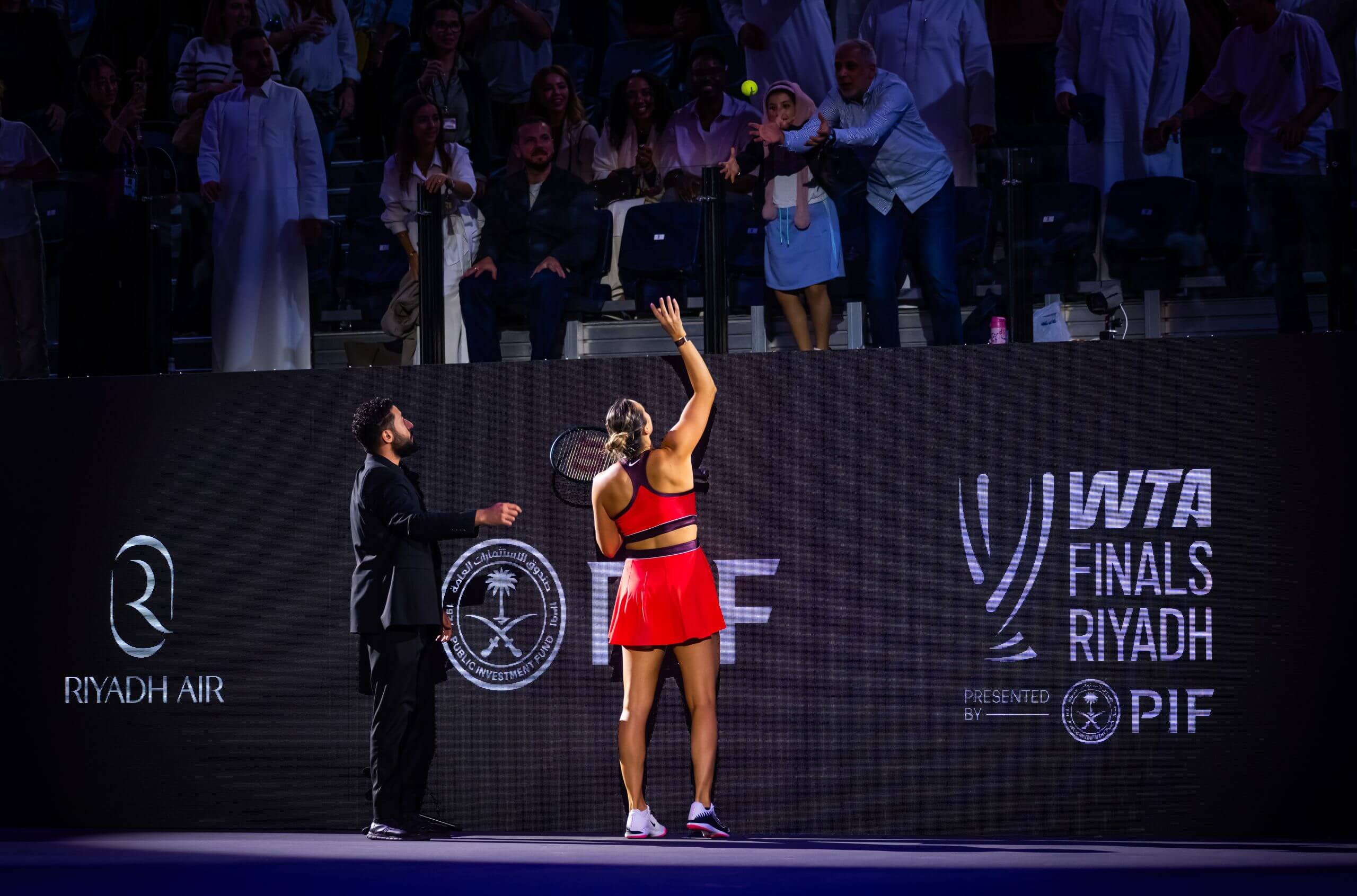Aryna Sabalenka throws a tennis ball to a woman in a stand raised above a tennis court, in front of hoardings advertising the Public Investment Fund and WTA Tour Finals Riyadh.