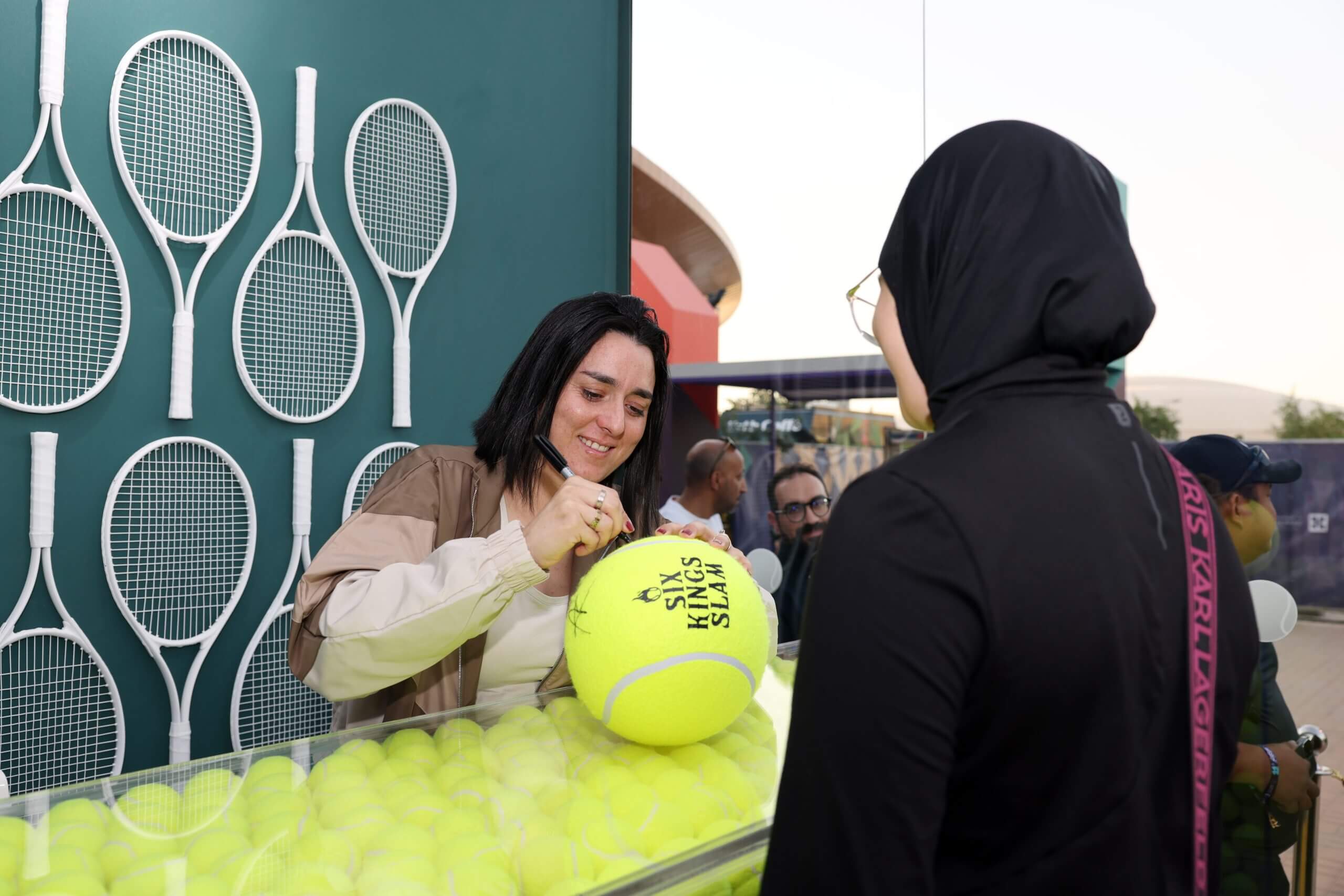 Ons Jabeur signs a giant tennis ball branded “Six Kings Slam” in front of a wall of white tennis rackets on a blue-green background, as a woman wearing a black outfit waits in front of her.