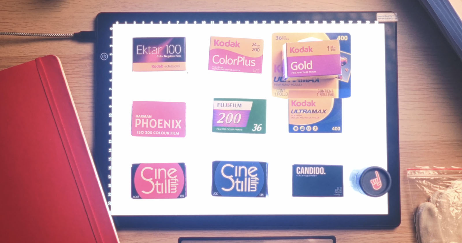 A lightbox displays various 35mm film boxes and labels, including Kodak Ektar 100, ColorPlus, Gold, Ultramax, Fujifilm 200, Ferrania P30, CineStill, and CANDIDO. A red notebook and plastic bag are nearby.