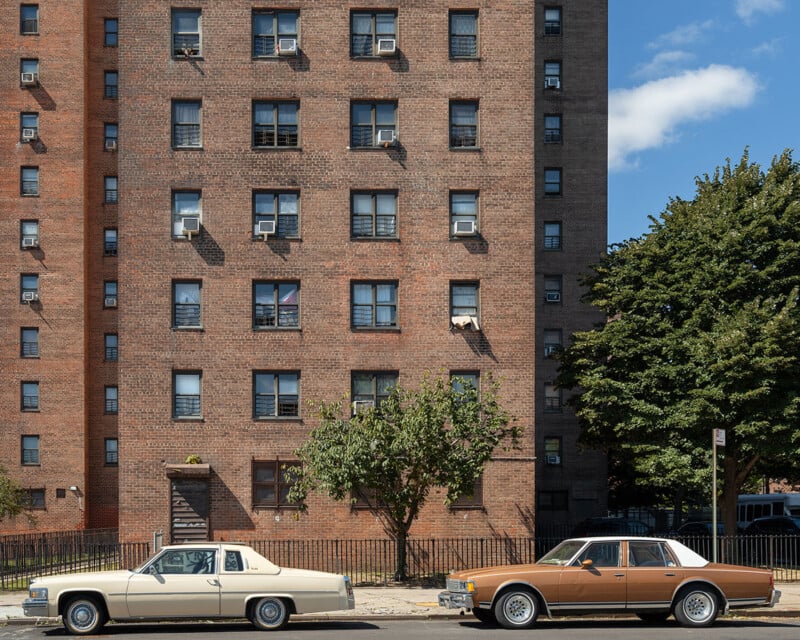 Two vintage cars, one cream and one brown, are parked on a street in front of a tall brick apartment building with many windows. Trees and a blue sky with some clouds are also visible.