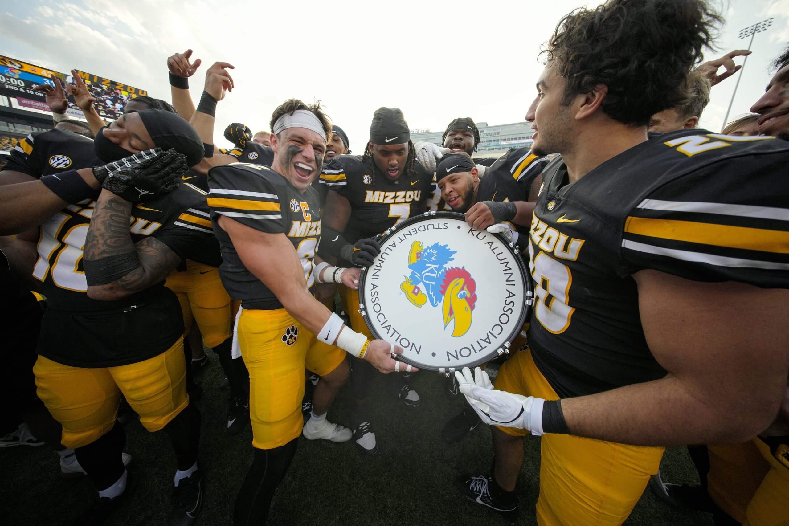 Missouri football players celebrate with the Indian War Drum trophy featuring a Kansas logo.