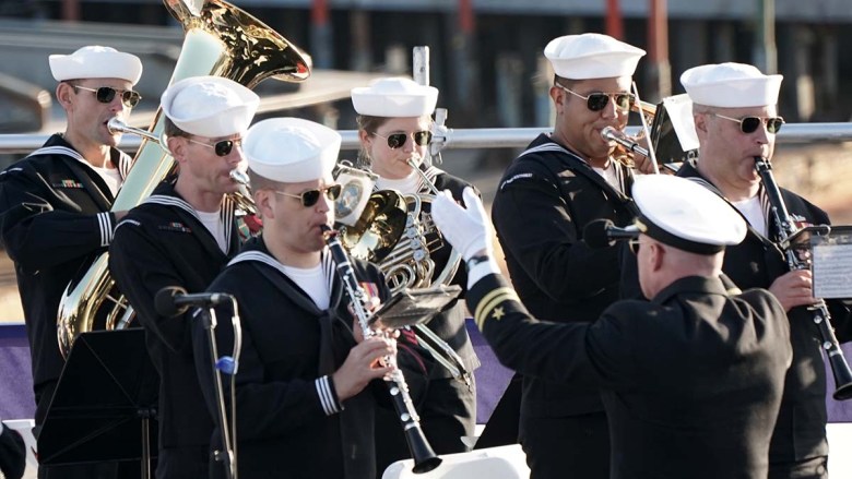 Musicians in black sailor uniforms play instruments, including horns, aboard a ship. They perform at community events, including Fleet Week.