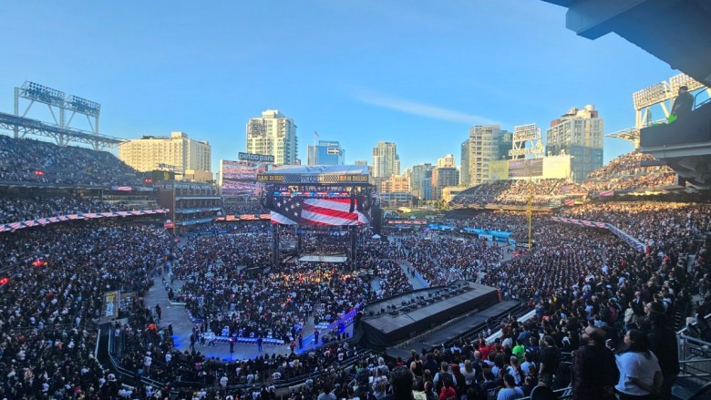A crowded stadium event at Petco Park with a central stage and large screen showing an American flag. High-rise buildings are visible in the background.