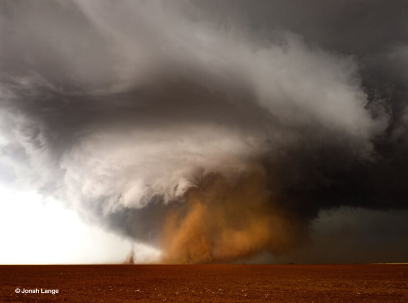 A massive, swirling storm cloud with a dense, dark funnel descends over a flat, reddish-brown landscape, kicking up dust and debris under a dramatic sky.