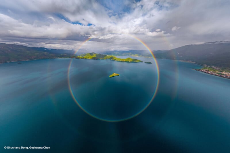 A wide, vivid circular rainbow arcs above a small green island in the middle of a deep blue lake, surrounded by mountains and cloudy skies.