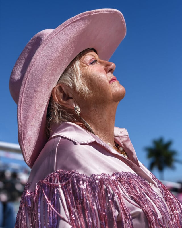 Older woman in a pink fringed jacket and wide-brim hat stands in profile under a clear blue sky, eyes closed and face lifted toward the sun; a blurred palm tree is in the background.