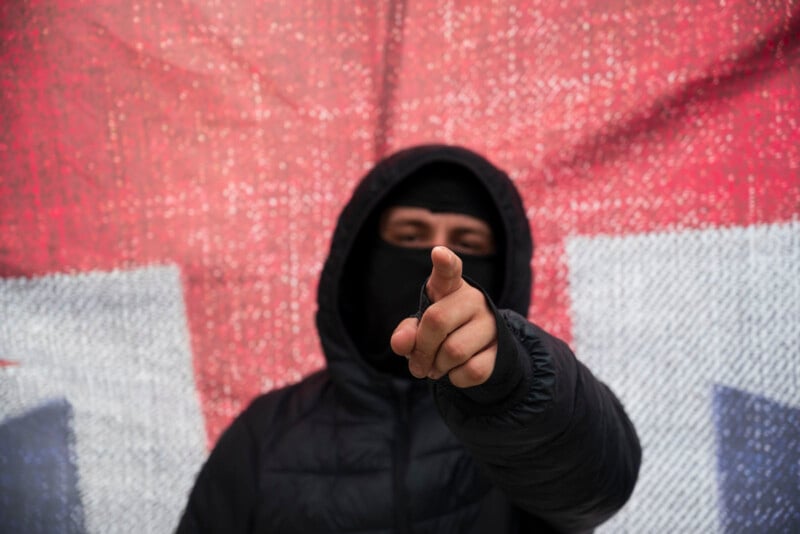 A person wearing a black hooded jacket and a black face mask points directly at the camera, standing in front of a large red and white textured backdrop.