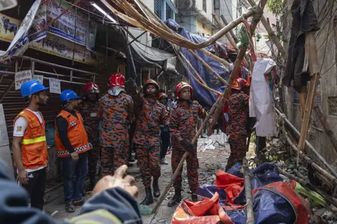Abdul Goni/Anadolu via Getty Images A group of emergency workers in mutlicoloured camouflage pattern outfits work to rebuild structures made of bamboo damaged in an alleyway in Dhaka.