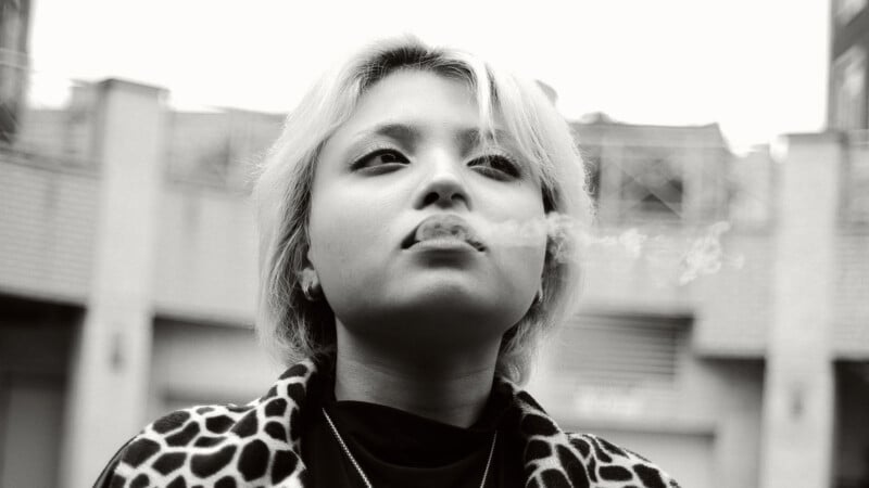 A person with short, light hair and a leopard print coat exhales smoke while looking upward, with buildings blurred in the background. The photo is in black and white.