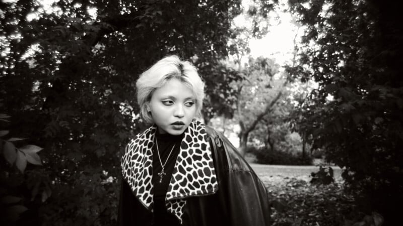 Person with short, light hair and a patterned scarf stands outdoors among trees, looking to the side with a thoughtful expression. The black and white photo creates a moody atmosphere.