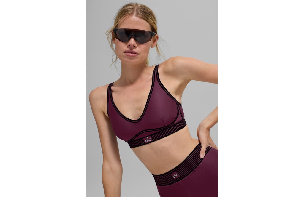 A model wearing a maroon Alo sports bra and leggings with a black elastic waistband and black sunglasses.