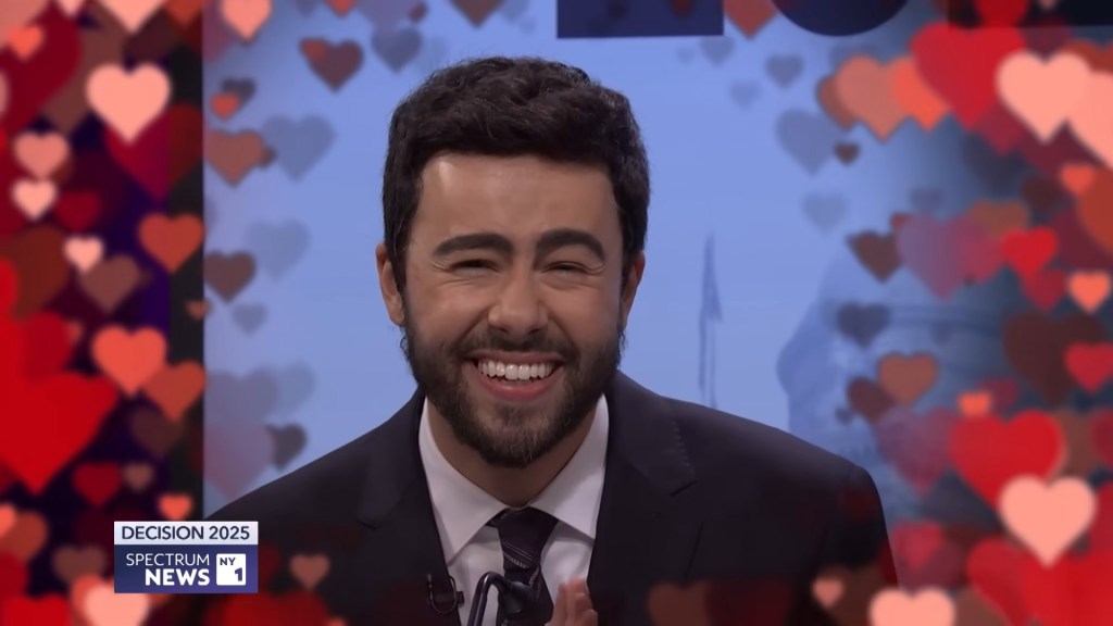Ramy Youssef as Zohran Mamdani laughing during a debate in a Saturday Night Live sketch.