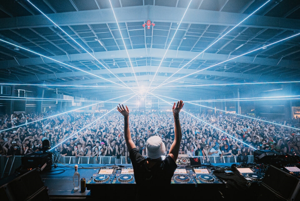A DJ with his arms raised stands onstage in front of a huge crowd.