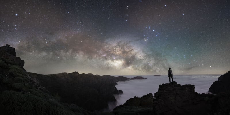 A person stands on a rocky cliff at night, overlooking a valley filled with clouds beneath a starry sky and a bright, colorful Milky Way stretching across the horizon.