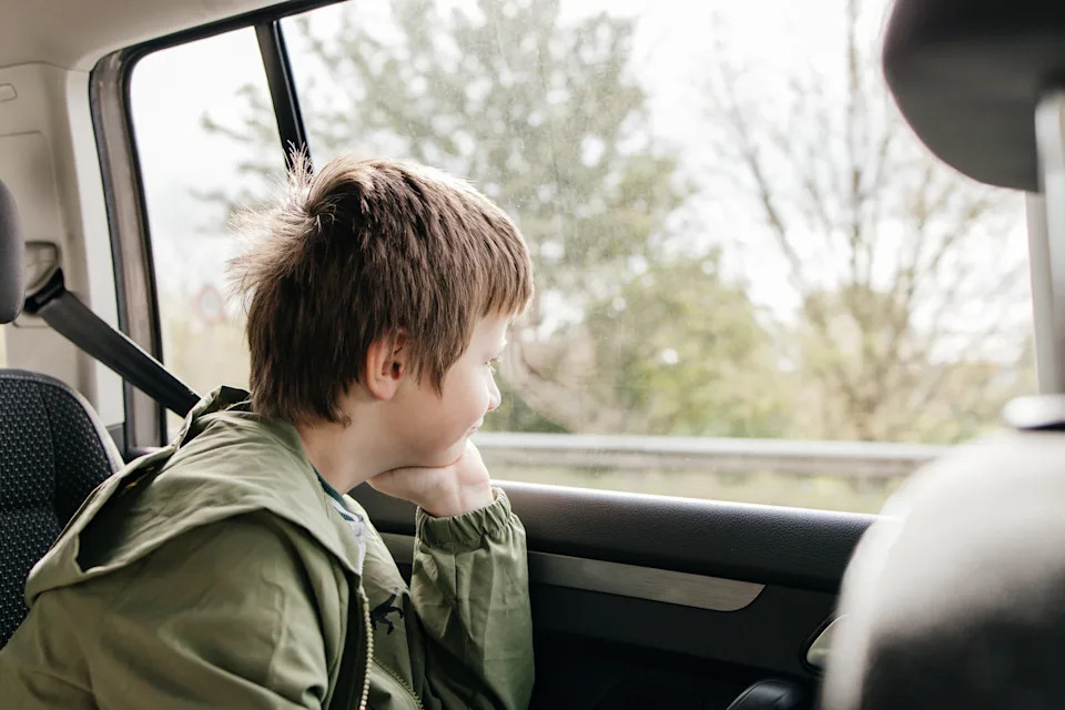 Child in a car looks out the window, resting chin on hand, wearing a casual jacket