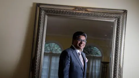 Toronto Star via Getty Images A photo of Deepak Paradkar from 2013. His reflection is photographed in a mirror with an ornate, silver border. He is looking at the mirror and smiling, wearing eye-glasses, a heathered grey suit with a pink tie and matching pocket square.