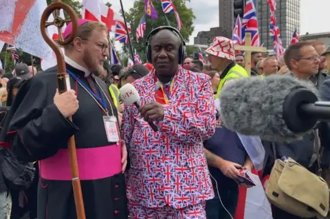 A bishop in religious clothing, holding a religious wooden stick, stands in the middle of a rally in central London with lots of St George's flags and Union Jacks in the background, while being interviewed by a man wearing a Union Jack themed blazer and trousers.