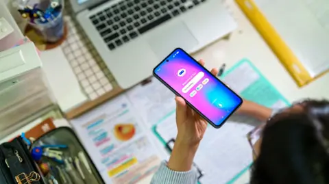 Getty Images A young female student seen from above interacts with an AI chatbot on a smartphone while studying at a desk with a laptop, notes and stationery. The scene highlights modern learning and technology integration.