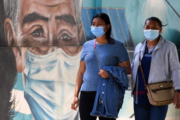 People wear surgical masks as they walk down an avenue in Tegucigalpa with a large painted mural of a masked face.