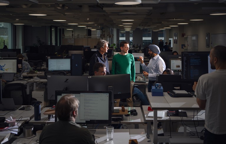 Many people work in a dark office. Three men are talking, standing between the desks in a better-lit area.