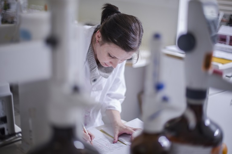 Woman taking notes in lab with lab equipment in the foreground and background.