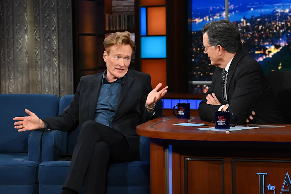Stephen Colbert and Conan O'Brien are having a lively conversation on a late-night set, seated with mugs on the desk