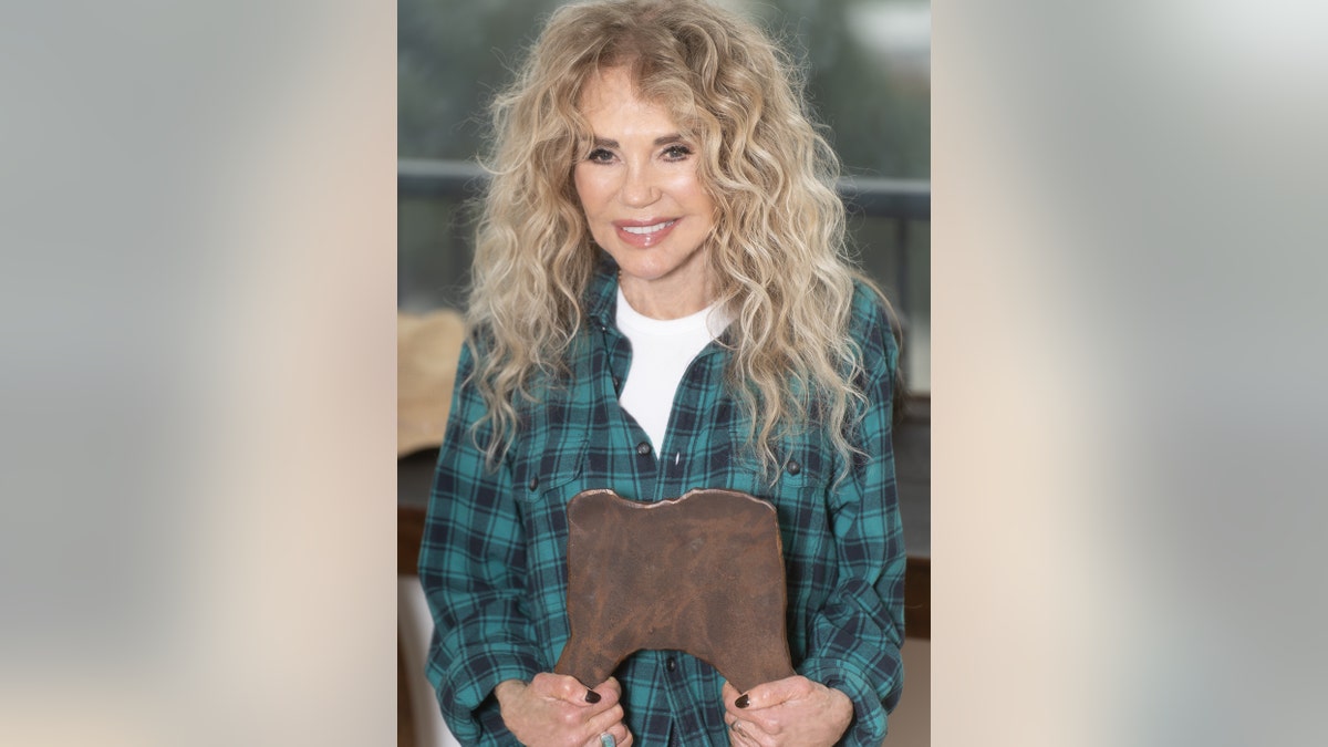 Dyan Cannon holding onto a wooden chair and smiling in a green plaid blouse with a white shirt.
