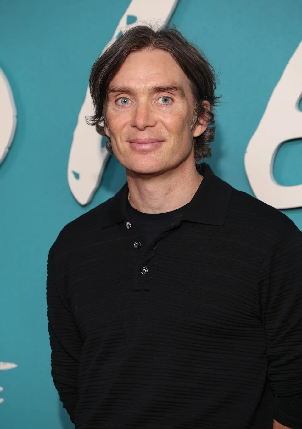 Person with medium-length hair wearing a black polo shirt, standing against a blue background with large white text
