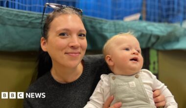 Leigh Walker and her baby Clyde - A woman with sunglasses on her head smiling at the camera. She's black hair in a ponytail and a black jumper. She is holding on her lap her baby who is wearing green dungarees and a white baby-gro. He's looking away from the camera and has a tuft of blonde hair on his wee head.