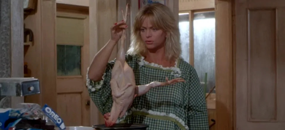 Person in a checkered dress holding a plucked chicken by its feet in a kitchen with a concerned expression